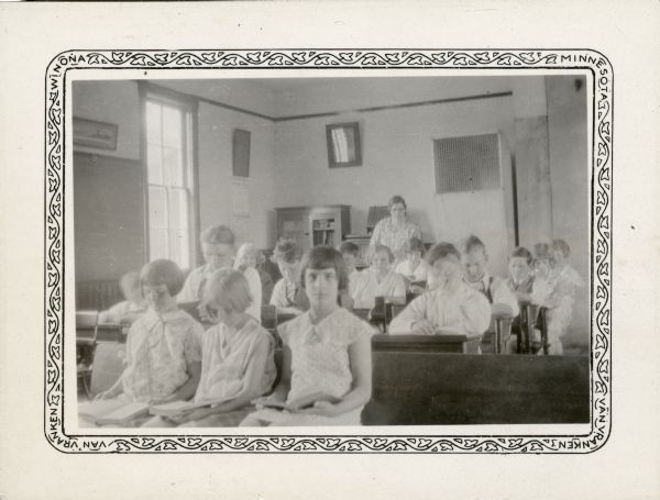 Students in the one-room Peck School read at their desks and sitting on a bench as their teacher, Mable Crosby, observes from the back of the room.