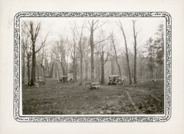 Two cars are parked in a grove of trees; people sit at a picnic table in the background. There are other tables in the area, as well as benches and a trash can.