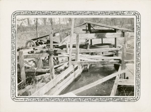 An open wooden structure built over a small mill race supports a vertical water wheel. A large belt runs horizontally from the water wheel to a line shaft and drives a generator. The generator provided electric power for the Teegarden farm.
