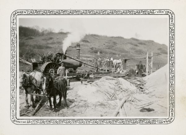 A steam tractor provides power for a portable sawmill owned by Jake Karken. The large sawblade is still as men pose for the photographer. There is a team of horses in the foreground near large piles of sawdust. Logs are stacked on the slope in the background. Karken's children attended the Peck School.