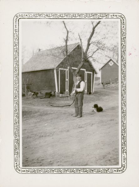 Fred Price poses in a farmyard holding a grain cradle which was made by his father and had been in the family for 55 years. A small granary and another outbuilding stand in the background. A dog lies on the ground behind Price.