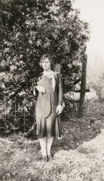 The teacher at the New Fane School, District No. 2, Town of Auburn, Fond du Lac County, poses near a fence with shrubbery behind her.