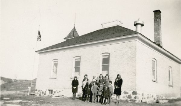 Thirteen students and their teacher, Marie Adams, pose at the side of the New Fane School. They are wearing coats and hats. The school is a singe-story brick structure with apparent bell tower and decorative widow's walk. There is a tall flag pole in front of the school.