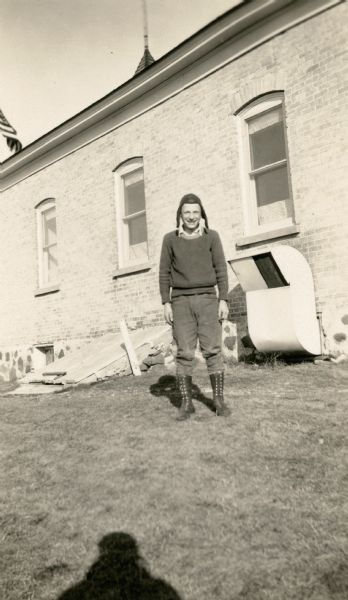 Martin Gutekunst, 14, wearing high boots, sweater, and leather hat, poses at the side of the New Fane School. He is identified on the photograph as the "leader of mathematics class". There is a large vent extending from a cellar window as well as a cellar door bulkhead.