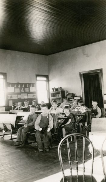 Students at the New Fane school pose in the classroom during lunch time. Some of the children hold cookies or sandwiches. On several of the desks there are lunch boxes and thermos bottles. The classroom ceiling is wood.