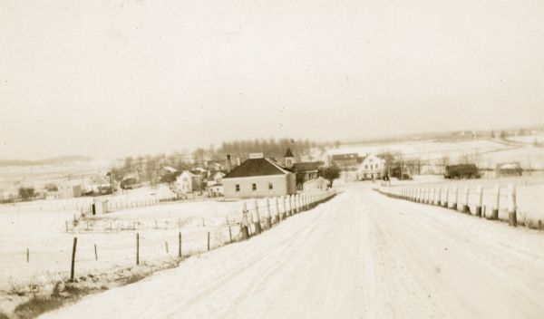 Looking west along County Trunk Highway SS (now CTH DD) from the hill east of the New Fane School. The brick school, with its bell tower and widow's walk, is on the left. An outhouse sits in the fenced school yard behind the school. The houses and businesses of New Fane are in the background. There are post and cable guardrails along the road. Light snow covers the ground.
