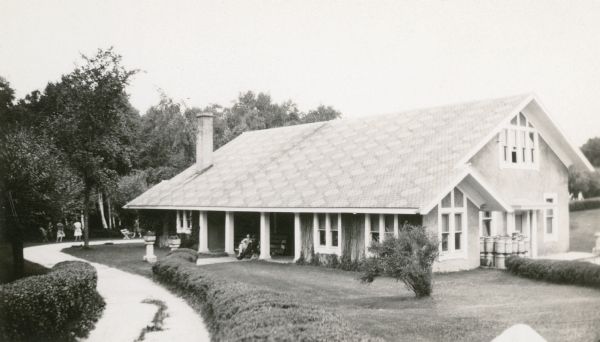 People relax on a bench on the front porch of the long, two-story stucco coldwater hatchery building on the grounds of the Fish Hatchery at Wild Rose. There is decorative half timbering in the gables, and a geometric design on the roof made by different colors of shingles. People are on the broad sidewalk along the left. Metal containers are stacked by the side door of the building. The grounds are well groomed, with decorative planters along the sidewalk which leads to the porch.