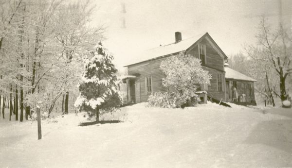 A one and one-half story house stands in a snowy landscape. A woman is walking toward the back door, hiding her face with a hand. The house was the childhood home of Marie E. Adams, the teacher at the New Fane School. At the time of the photograph the house belonged to Nellie L. White.