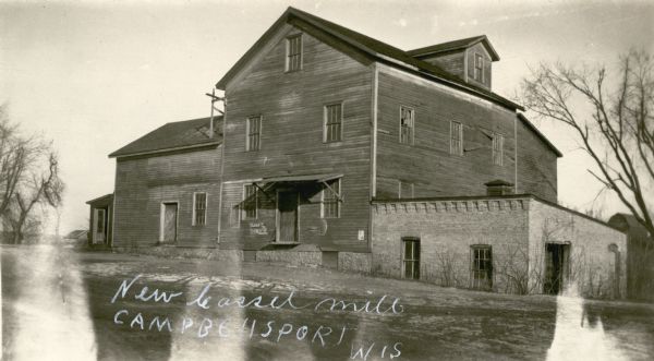 A large wood frame mill building with one-story brick addition. There is an elevated door with a roof which was probably used for deliveries. In the far background on the right is a barn.