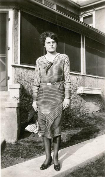 Marian Metcalf, teacher at Dyer School, poses on a sidewalk in front of the porch of a stuccoed house.