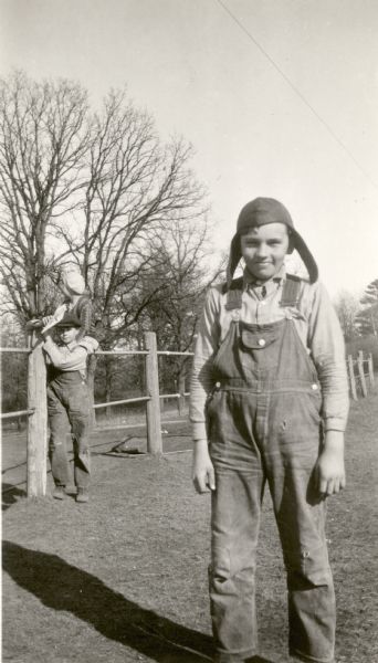 Claire Alexander, 12, identified on the reverse of the photograph as "Boy Champion of 4-H Club" posing wearing bib overalls and a cap with earflaps. Two other students are seen in the background resting on a fence made from wooden posts and metal pipe. One boy appears to be holding a paper airplane. Alexander's parents are identified as Gorham Alexander and Erna Alexander, deceased.
