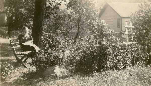 A woman sits and reads on a wooden slat bench in a flower garden. There are ornamental plants in a wash tub and other containers; a rose bush nearly covers a trellis. There is a small frame building in the background. On the reverse of the photograph is written, "A flower garden partly maintained by Wilma Wetter age 9. Native of Lancaster, Wisconsin. Rueben Wetter is owner of the farm."
