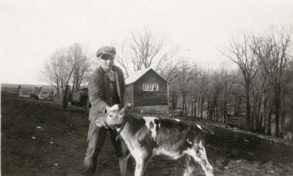 Claire Alexander posing with a young calf in a pasture near a wooded ravine at his father's farm. There is a manure spreader and shed in the background.