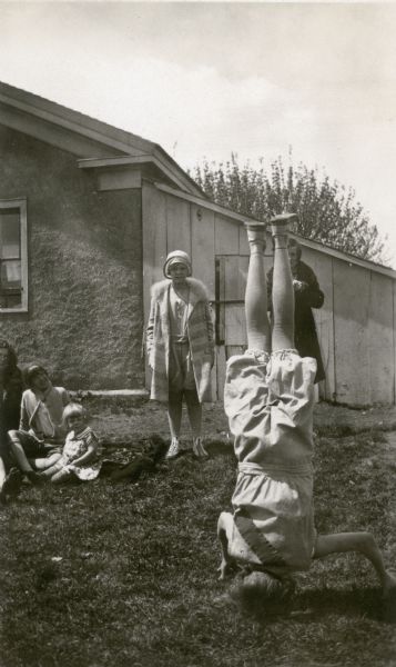 A girl wearing bloomers and long stockings performs a headstand as other girls look on. One of the bystanders is also wearing athletic clothes under her coat.