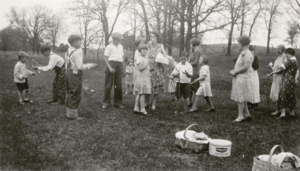 Teacher Marian Violet Metcalf, center, stands among her students as they prepare to roast marshmallows at the school picnic. Picnic baskets and a large marshmallow can rest on the ground. There are several women on the right. The picnic was held in a pasture owned by Sherman Pittenger, just down the hill from the Dyer School.