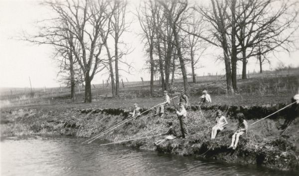 View across water towards a group of pupils from the Dyer School fishing from the banks of a stream "about a mile south of school." They are using long cane poles.