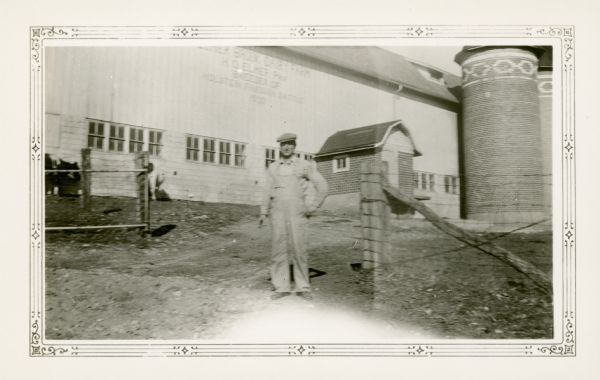 Henry Otto Elmer, wearing a cap and bib overalls, poses in front of his large dairy barn. There are twin silos on the right with a decorative pattern in the clay tiles. The sign on the barn identifies the Elmer Brook Dairy Farm, H.O. Elmer, Prop., Breeder of Holstein Friesian Cattle, 1920. Mr. Elmer was the clerk of the Farmer's Grove School District.