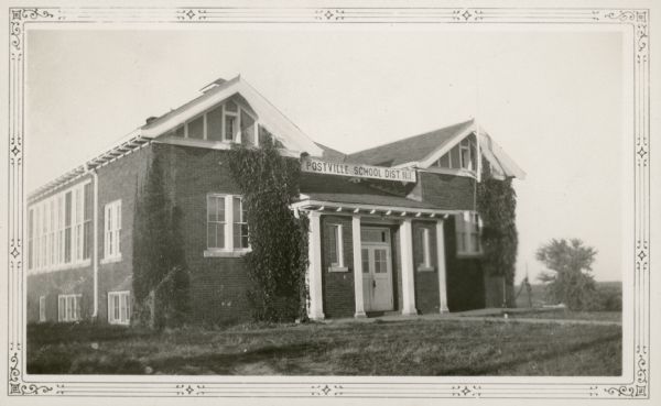 A prominent sign identifies the Postville School, District No. 1. The school is brick, with twin gables in the front; vines grow up the walls. The gables have decorative half-timbering with stucco infill. The bell tower is just visible at the peak of the roof. A note on the reverse of the photograph states that the school had an average enrollment of 40 pupils.