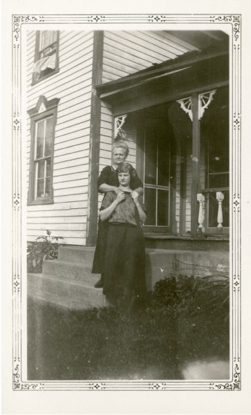 Selma Eismoe, the teacher at Farmer's Grove School, poses with her mother, Mrs. H.O. Eidsmoe. Mrs. Eidsmoe stands on concrete porch steps. A curtain blows from an upstairs window.