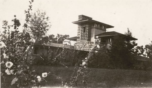 Exterior view of Frank Lloyd Wright's residence, Taliesin, from the southeast, emphasizing the broad overhangs and horizontal lines of the structure. Hollyhocks bloom in the foreground. This photograph was submitted to the Wisconsin Rural Schools Survey by Selma Eidsmoe, teacher at Farmer's Grove School in Green County, as her favorite place to visit.