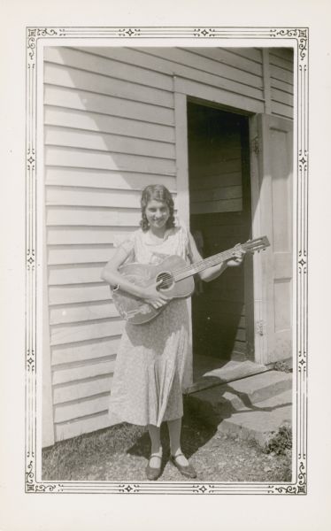 Arlene Elmer, 14, poses outside the Farmer's Grove School with a guitar. On the reverse of the photograph is written "This pupil is leader in plays and games as well as in art and music."