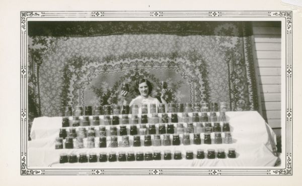 Esther Elmer, 20, poses with jars of fruits and vegetables which she has canned. A fancy carpet is hanging behind her as a backdrop. She wrote on the reverse of the photograph: "I have exhibited 4 years in canning projects and won 35 firsts and 51 seconds. I entered some fruit and vegetable jars in the National Canning Contest at the Century of Progress at Chicago (1933) and received special prizes."