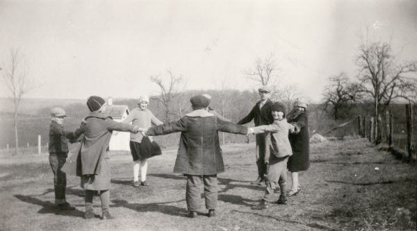 Children have clasped hands and formed a circle in a game of "Drop the Handkerchief." They are in the schoolyard of Farmer's Grove School, District No. 3, Town of York and New Glarus. There is an outhouse with privacy fence in the background.