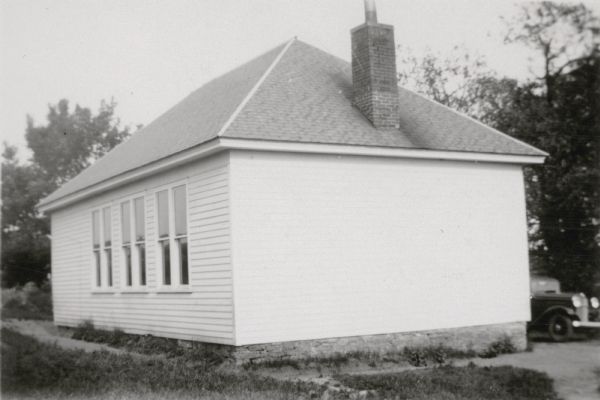 A view, from the rear, of the small, wood frame Strawberry School, District No. 4. The school has a hip roof and brick chimney. A car is parked beside the far side of the building. On the reverse of the photograph is written "Located on Highway 80, 3 miles north of Highland Wis."