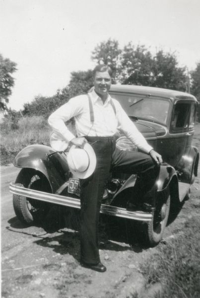 Orin Swingle, teacher at Strawberry School, poses with his left foot on the bumper of an automobile. He wears suspenders and holds his hat in his right hand.
