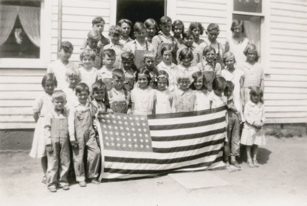 The students at Strawberry School pose in front of the school building. The youngest students, in front, hold an American flag. There are curtains and a paper flower in the window. The boys all wear bib overalls.