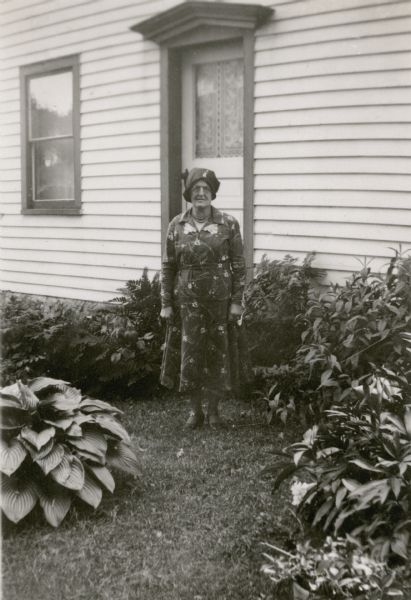 Bertha Swingle, wearing a print dress and hat, poses outdoors in front of a door at her home. There is a large hosta plant on the left and other shrubbery in the yard. Mrs. Swingle's son Orin was the teacher at Strawberry School, District No. 4.