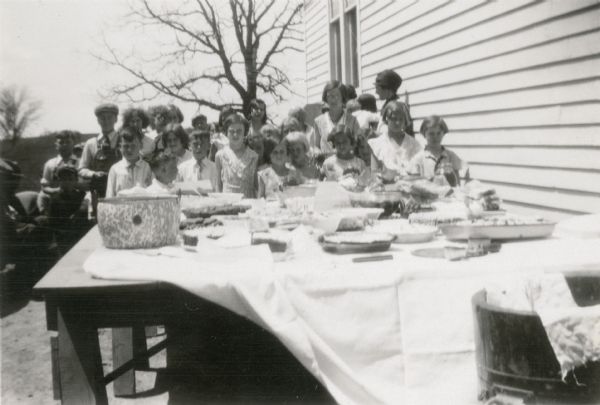 Pupils at Strawberry School pose behind a table laden with food brought for the "last day of school" picnic. The children wear "good" clothes; two boys with bib overalls wear neckties.