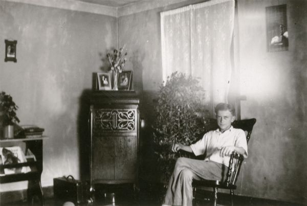Leland Kopps, a student at Strawberry School, sits in a chair near a Victrola-style cabinet phonograph in his farm home. There is a large potted plant in front of the window.