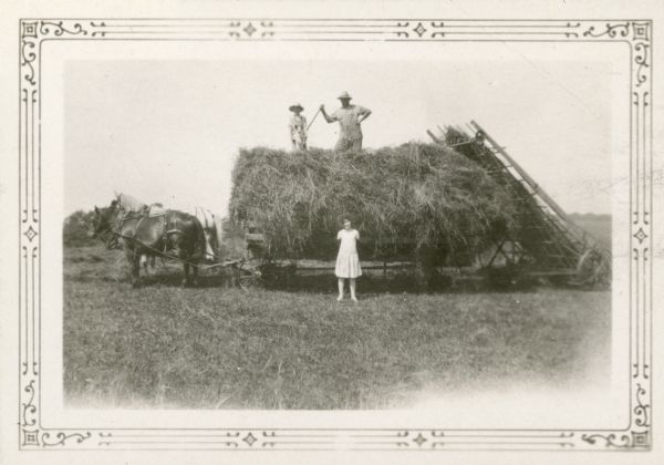 Leland Kopps holds a dog as he poses standing with his father John on top of a load of hay. A team of two horses are harnessed to the wagon which pulls a hay loader. Lillian Kopps stands on the ground in front. Leland and Lillian were students at Strawberry School.
