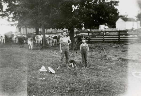 Russell, left, and Ira Wepking pose as they pause from bringing in the dairy cows; they have taken off their hats, which are on the ground. A small dog stands between them.