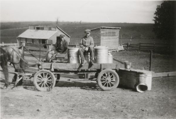 John Hurda, left, and Russell Wepking pose with milk cans on a farm wagon. Hurda holds the reins to a team of two horses. There are two small sheds behind them and fields beyond.