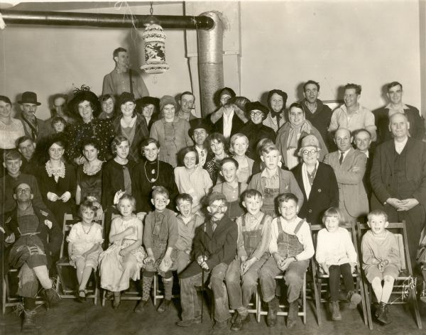 Pupils of the Northey School, District No. 7, pose with their parents at a Halloween "Hard-time" party at the school. Many in the group wear ragged or outdated clothes and mismatched stockings. A paper Japanese lantern with a witch design hangs from a light fixture. On the reverse of the photograph is written, "Pupils entertained their parents at a Hard-time party on Hallowe'en. Prizes were given for the best, or rather worst, costumes. An old fashioned spell down was held."