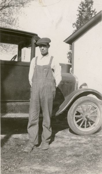 William Richard Leonard, the director of the Northey School board, poses next to an automobile. He is wearing bib overalls and a cap.