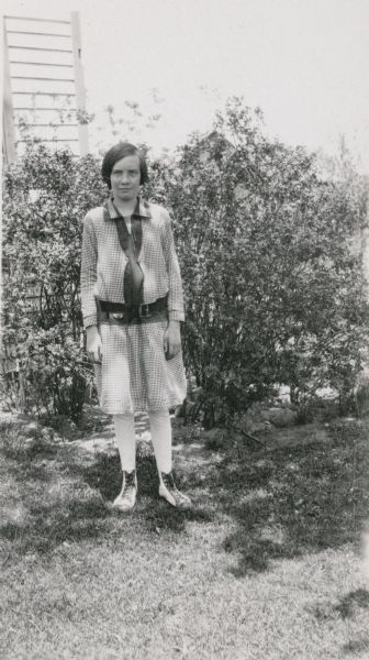 Edna Leonard, a student at Northey School, posing in front of shrubs near a building. On the reverse of the photograph is written: "Belongs to a local 4-H sewing club. She has won several prizes exhibiting garments she has [made]."