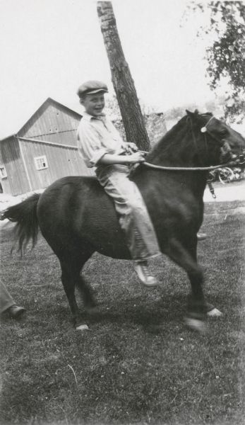 Richard Northey, 11, a student at the Northey School, poses on a pony. There is a barn in the background. On the reverse of the photograph is written "He has a fine Guernsey heifer calf in the [Jefferson] Co. Guernsey Way Calf Club."