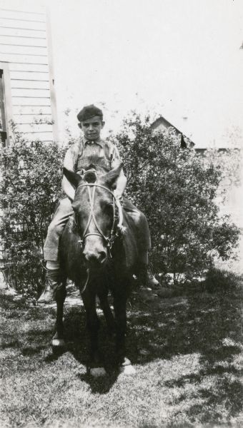 Harlow Hebbe, 10, a student at Northey School, poses on a pony in front of bushes at the side of a building. On the reverse of the photograph is written "Makes particularly interesting designs — such as are appropriate for posters and book covers."