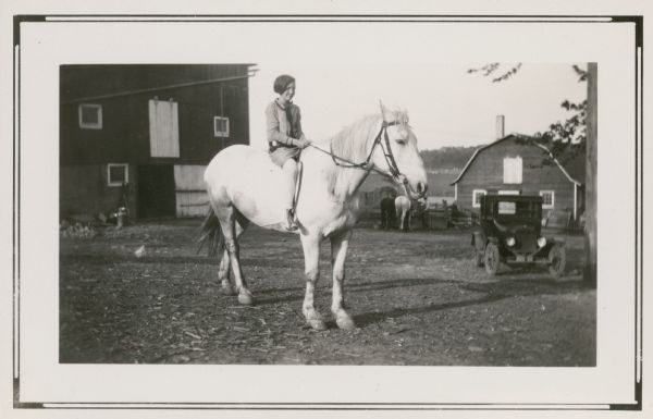Edna Leonard, a student at Northey School, poses on a horse on the Fred Leonard farm. There is an automobile and other horses in the background, as well as two barns.