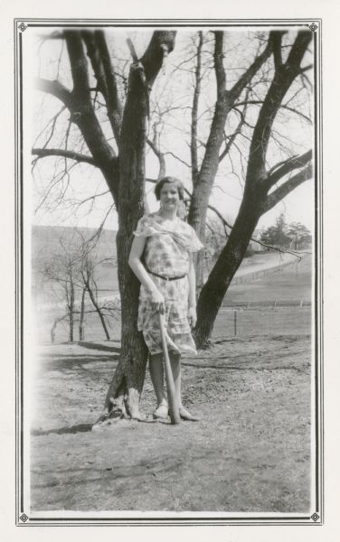 Esther Peterson, a student at Mud Branch School, District No. 2, poses leaning against a tree on a hill while holding a baseball bat. There is a road and farmhouse in the background. On the reverse of the photograph, Esther is identified as "Leader of highest class in English."