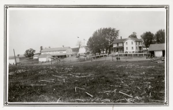 The Theodore Vinger farmstead sits on gently rolling terrain. A painted sign on the large barn identifies the owner. At the time of the photograph, Martin Peterson rented the farm. In addition to the large barn, there is a smaller barn and several outbuildings. A car is parked in front of a garage near the wood frame farmhouse. There is a windmill on the hill behind the buildings.