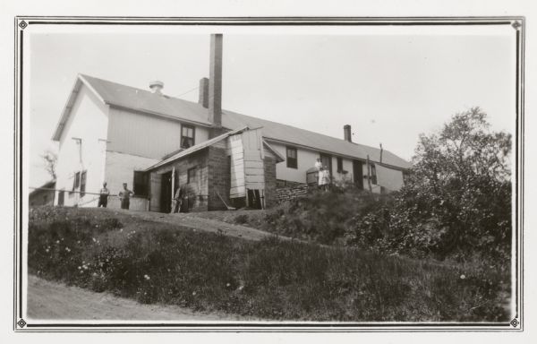 View up road towards two men posing near the front of a cheese factory on a small hill; on the right a woman and two girls stand near a side door. Typical of cheese factories, the building, which has three chimneys and a ventilator, is built close to the road and has living quarters behind.  A small addition made of clay tiles extends from the side. Flint Vinger was the owner and cheesemaker at the time of this photograph. On the reverse of the photograph he was described as "the best cheesemaker in this large area. His two daughters help watching and washing whey barrels." It is noted that he made Swiss, block, brick, Limburger and American cheese.