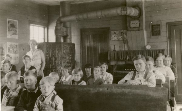Teacher Marjorie Carey stands behind her pupils sitting at their desks at the Mud Branch School, District No. 2, as they pose for their photograph. There is a large stove with ornate metal work in a corner of the room behind the teacher, and a parlor organ along the rear wall, centered between two doors. Two sets of twins are seated on the right; they are Glen and Gilman Blomquist and Viola and Violet Jacobson. A clock on a shelf records the time: 9:35.