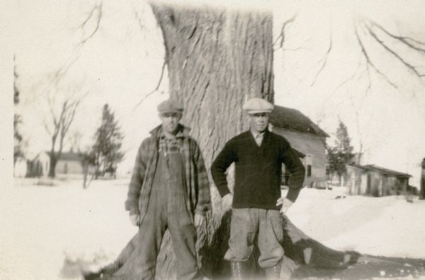 Lavaughn Cooper, in bib overalls and plaid jacket, and Bud Briggs, wearing knickers and a sweater, school board members of the Eagle Corners School, District No. 3, Township of Eagle, pose in front of a large tree. There is snow on the ground. There is a house and outbuildings in the background.