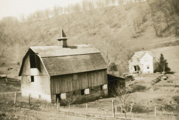 View down hill towards a barn on the Magnus Hofseth farm. Two cars are parked near the house. The barn has a gambrel roof and wooden ventilator. The loft door is open. A steep wooded hill rises behind the farm.
