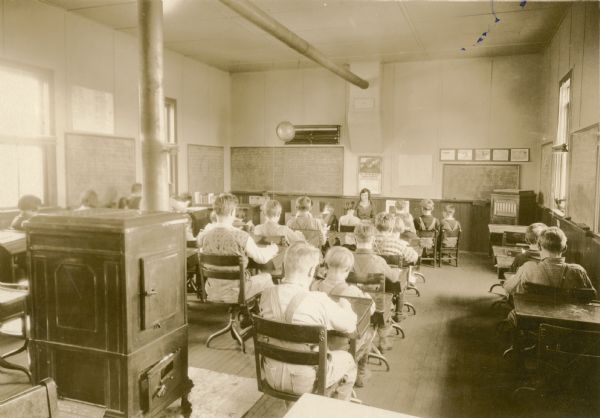 Glady Hofseth, the teacher at Eagle Corners School, sits with a small group of students at the front of the classroom while the other students work at their desks. There is a large stove in the back of the room. A Victrola style phonograph stands in a front corner.
