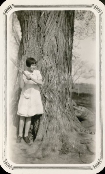 Irene Mervin, a 12-year-old student at Eagle Corners School, poses with a baseball bat in front of a large tree. On the reverse of the photograph is written "Irene is very good natured."
